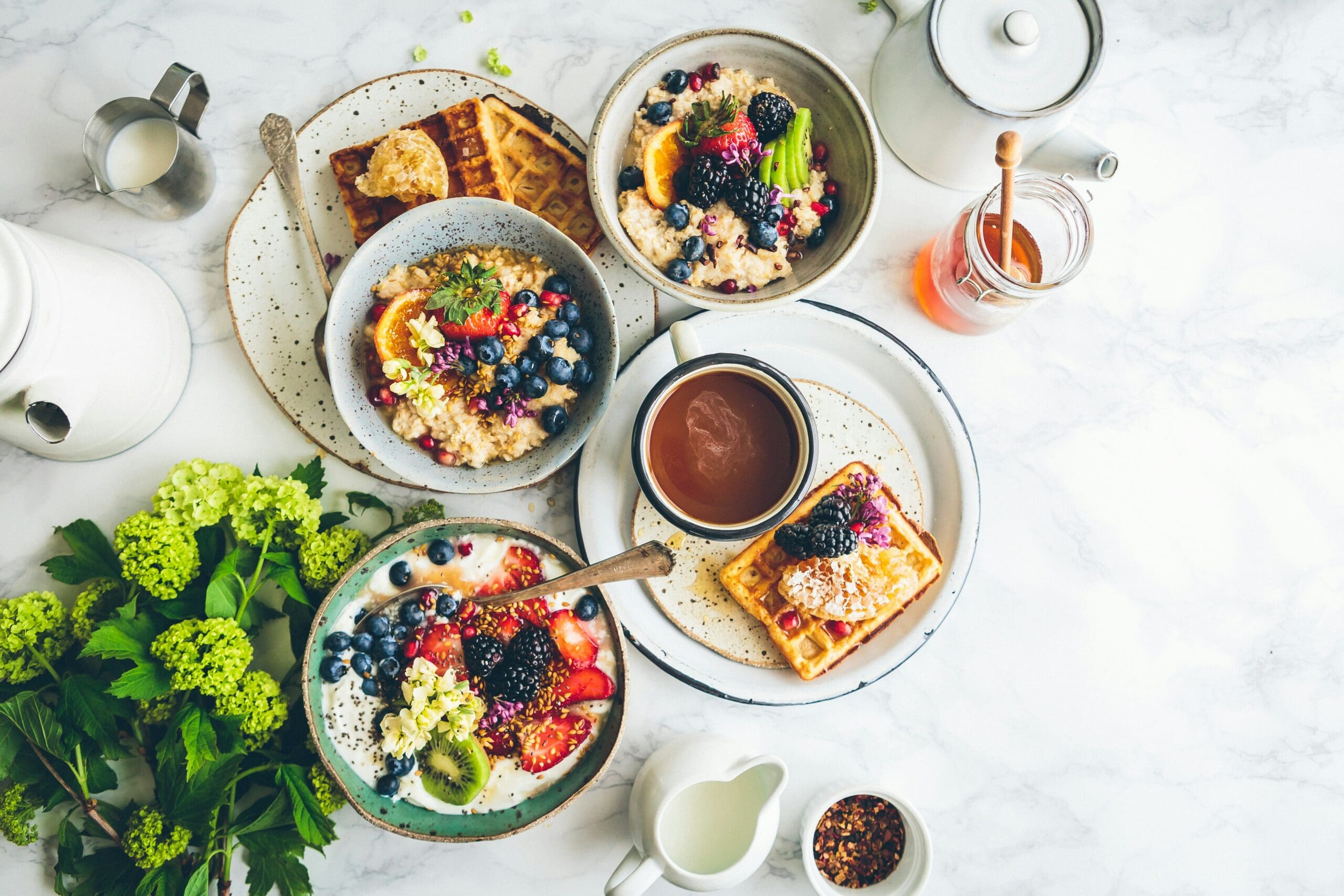 Breakfast spread with fruit-topped porridge and yoghurt bowls, waffles, tea and honey on a marble table.