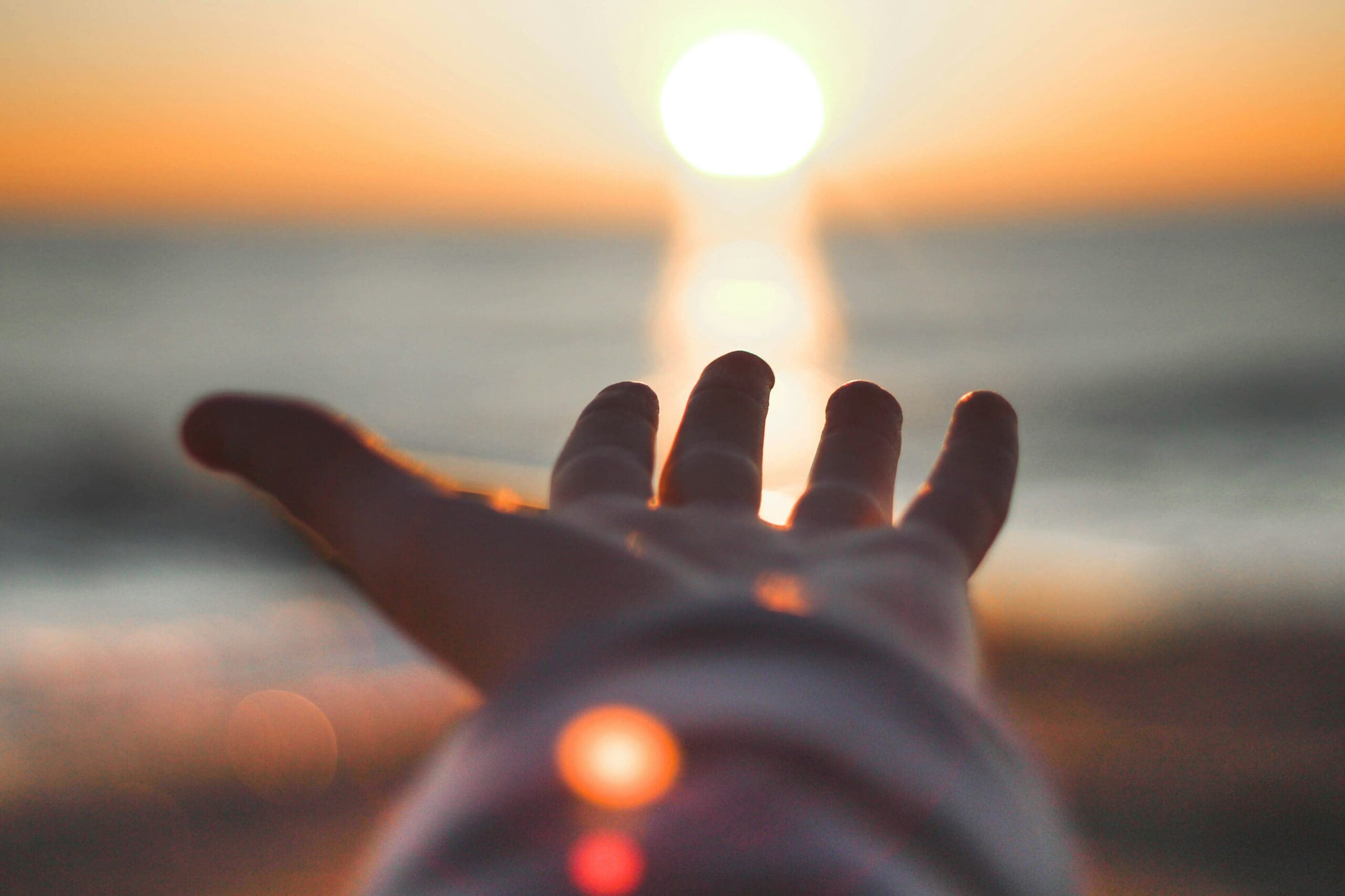 An outstretched hand reaching towards the sun over the ocean at sunset.